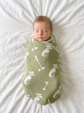 A sleeping newborn baby swaddled in an olive green blanket with a white golf ball and club pattern, lying on a white textured sheet.