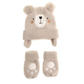 A baby knit hat and matching booties set in a light beige color, featuring a bear face design on the hat with pom-poms and ear flaps, and paw-print details on the booties.