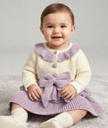 A baby sitting on a light gray carpet wearing a cream knit cardigan with a lavender ruffled collar and matching lavender bow skirt.