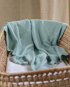 Light bgreen knitted blanket draped over a wicker chair with a neutral background