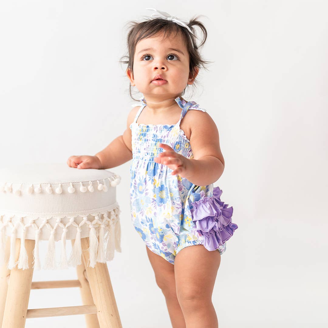 Baby wearing a floral romper with ruffles, standing next to a white stool.