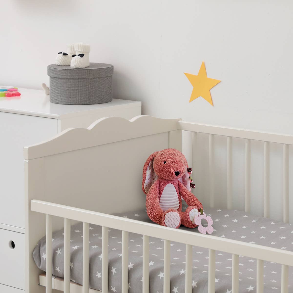 White crib with pink plush toy and gray star-patterned mattress cover in a nursery setting.