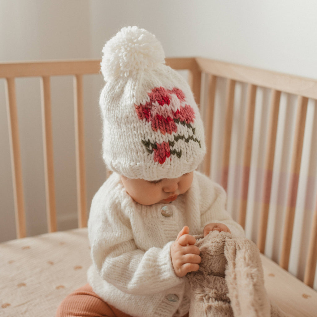 A baby sitting in a wooden crib wearing a cream knit pom-pom beanie with a red and green floral pattern, a cream knit cardigan, and holding a plush bunny toy.