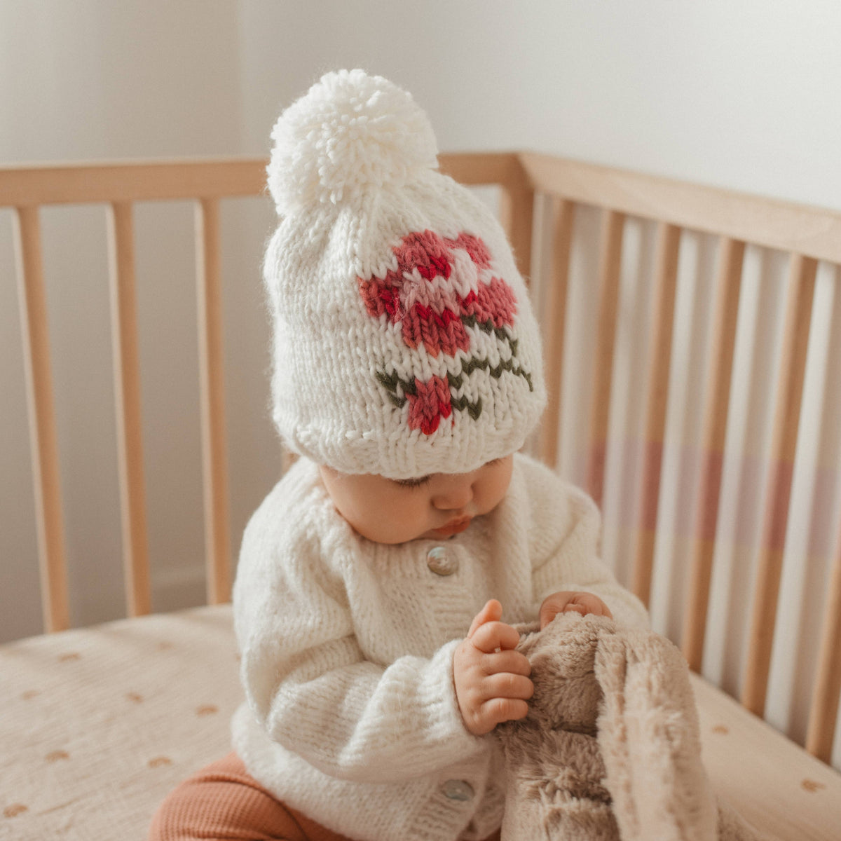 A baby sitting in a wooden crib wearing a cream knit pom-pom beanie with a red and green floral pattern, a cream knit cardigan, and holding a plush bunny toy.