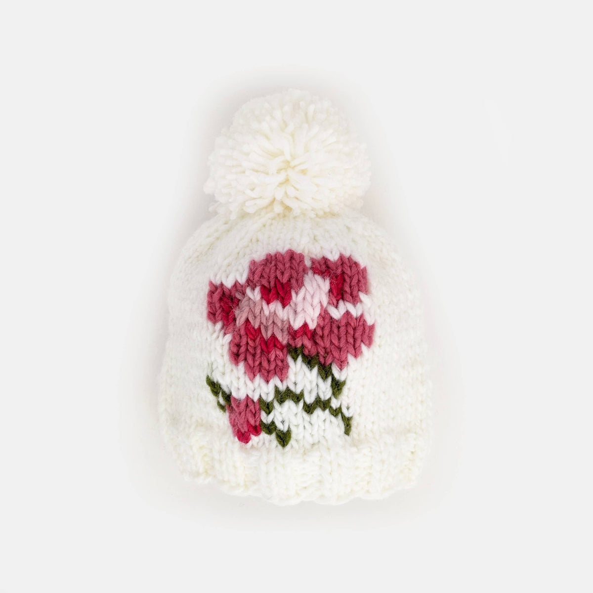 A white knit beanie with a fluffy white pom-pom and a knitted floral pattern in pink and green on the front, shown against a plain white background. AdoraBaby.co.uk