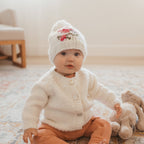 Baby wearing a white knit cardigan and hat, sitting on a carpeted floor with a teddy bear.