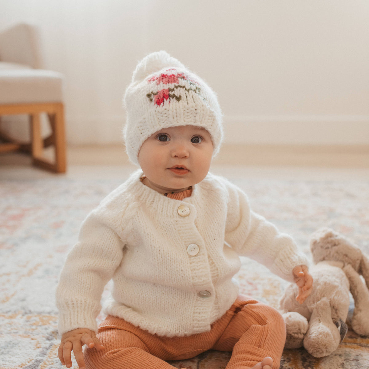 Baby wearing a white knit cardigan and hat, sitting on a carpeted floor with a teddy bear.