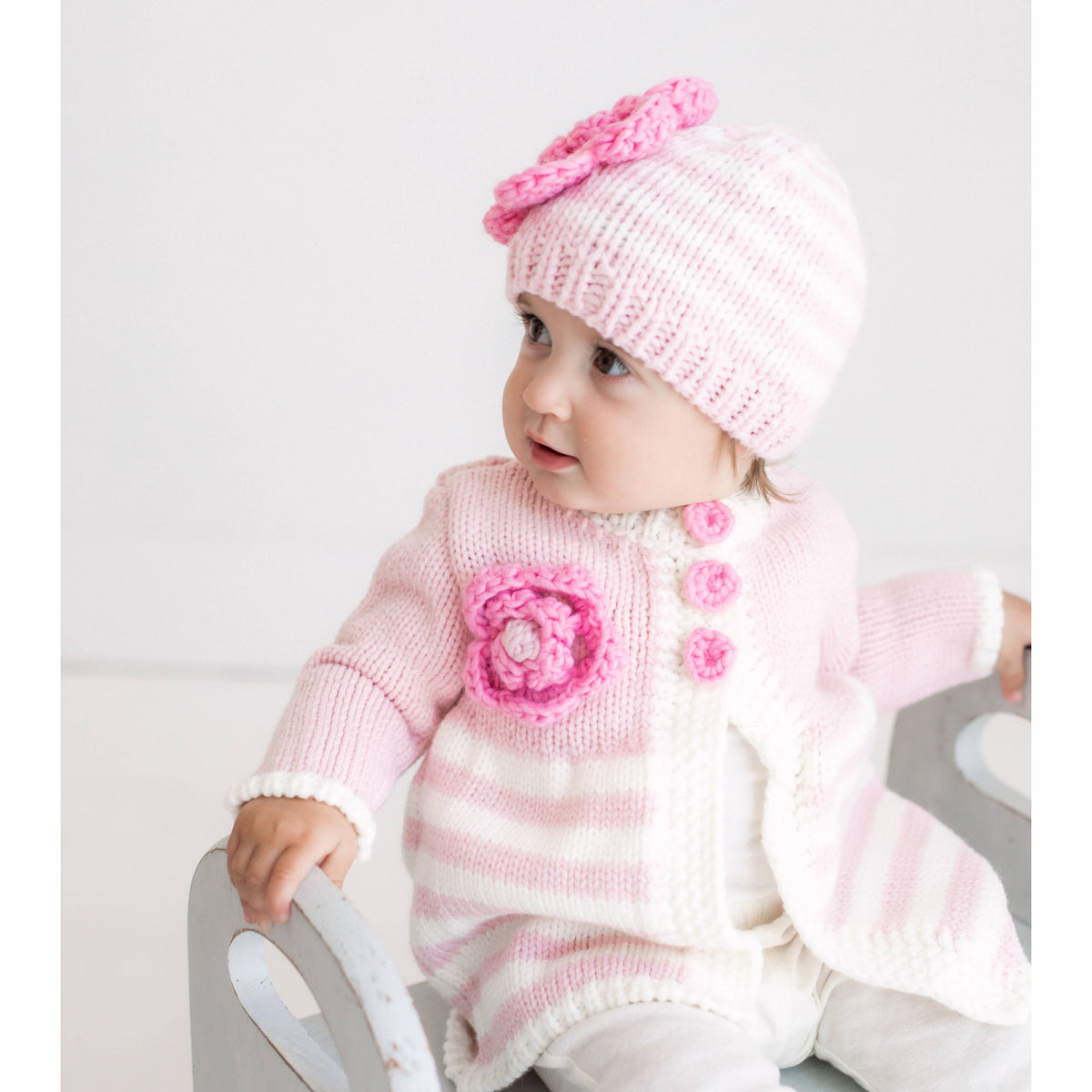 Baby wearing a pink knitted outfit with a matching hat on a light background