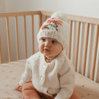 A baby sitting in a wooden crib wearing a cream knit beanie with a coral and green floral pattern, a cream knit cardigan, and peach pants.
