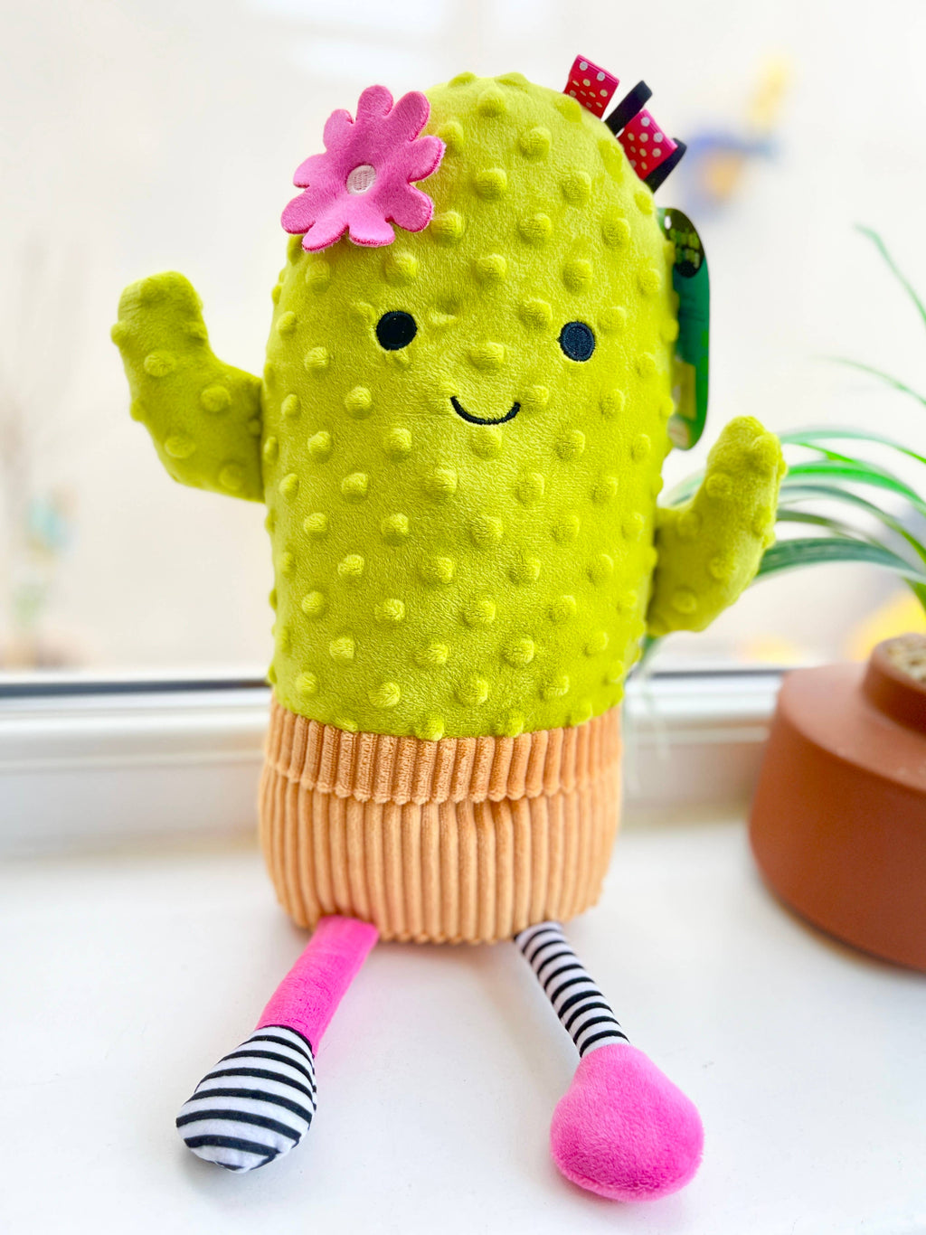 A cute cactus plush toy with a bright yellow-green textured body, orange corduroy skirt, pink and black-and-white striped legs, a pink flower on its head, and a red bow, sitting on a white surface.
