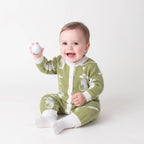 A smiling baby sitting on a light gray studio background, wearing a green and white golf-themed footed sleeper and holding a white golf ball.