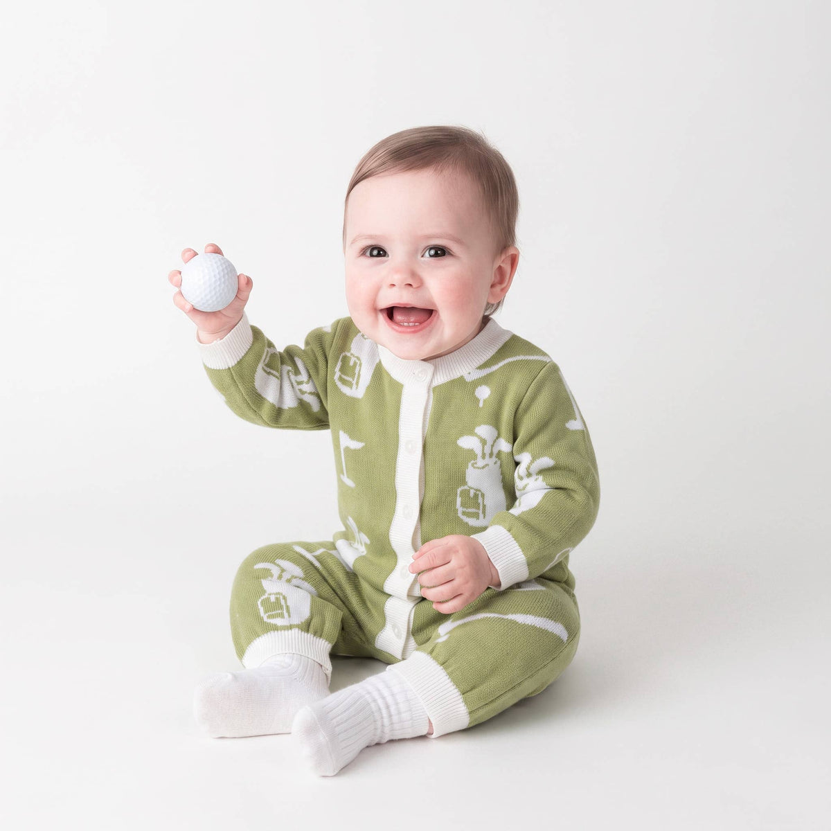 A smiling baby sitting on a light gray studio background, wearing a green and white golf-themed footed sleeper and holding a white golf ball.