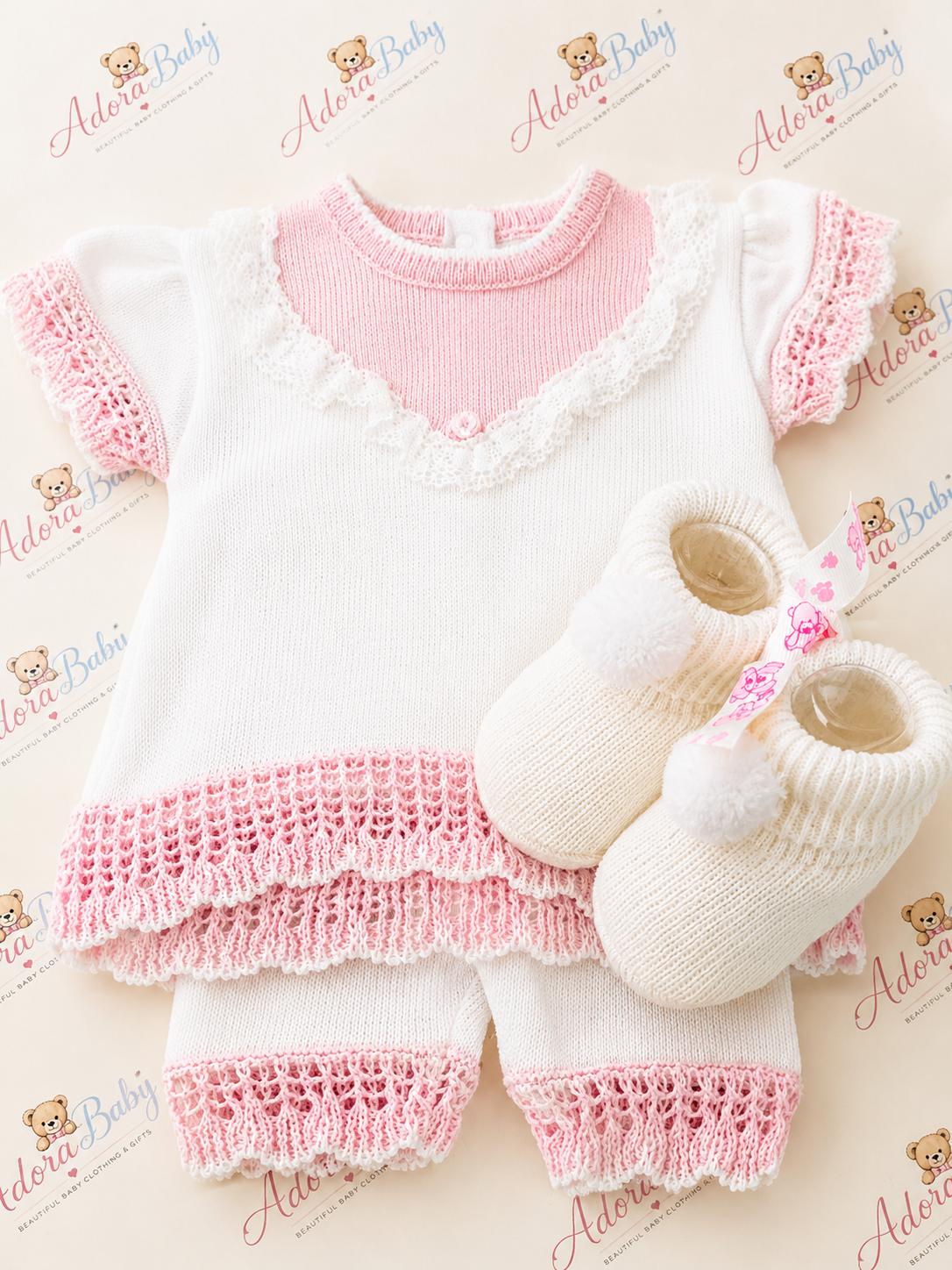 Adorababy.co.uk  baby knit outfit set featuring a short-sleeve top and matching pants in white with pink ruffle trim, accompanied by cream booties with pink floral accents, laid flat on a patterned background.