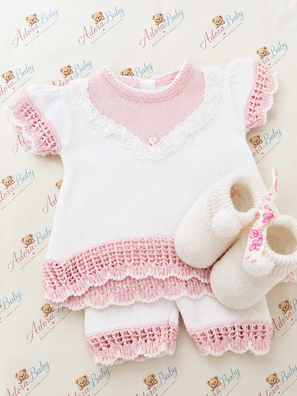 A baby clothing set laid flat on a branded cream background, featuring a short-sleeve romper in white with pink trim and lace, matching white shorts with pink lace trim, and a pair of white knit booties with pink floral embroidery.