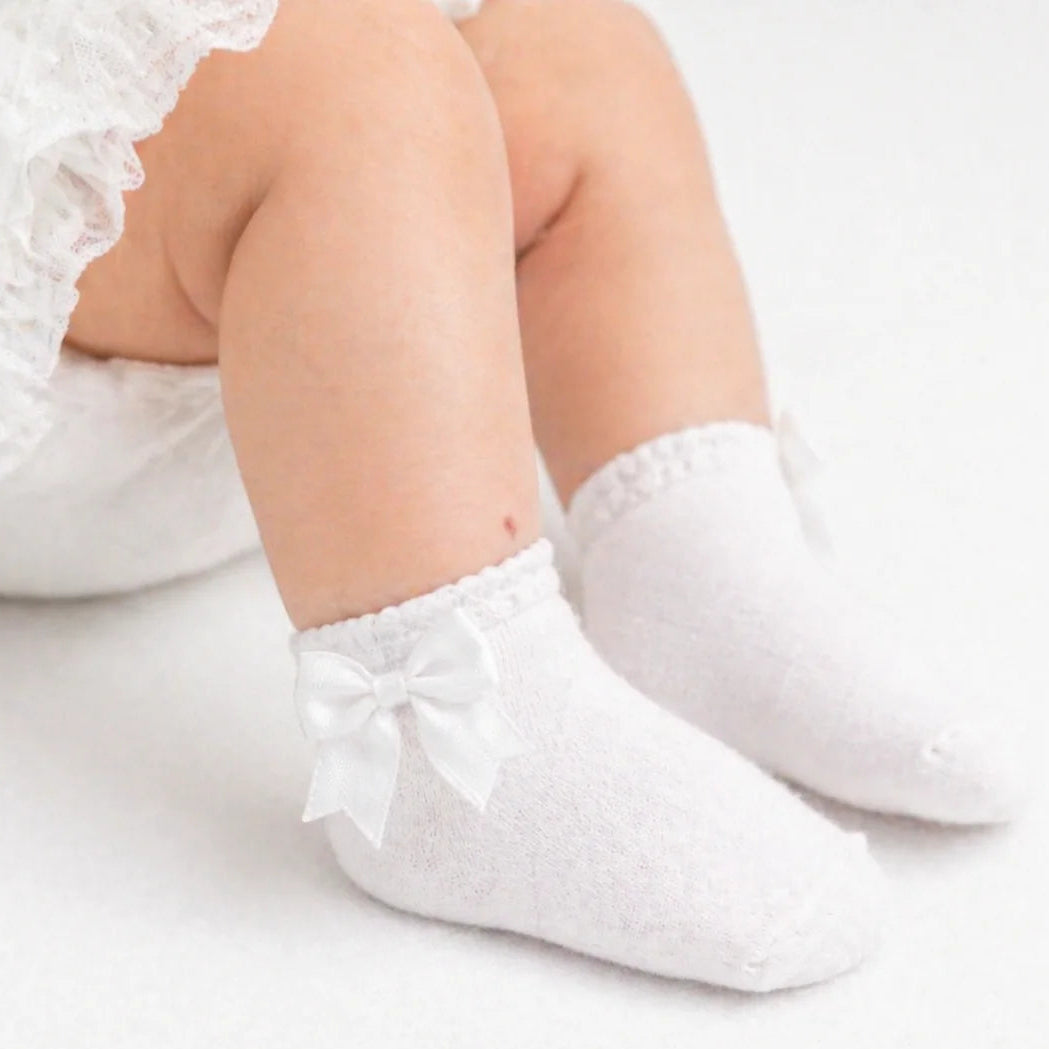 White baby socks with bows worn by a child on a light background adorababyco.uk