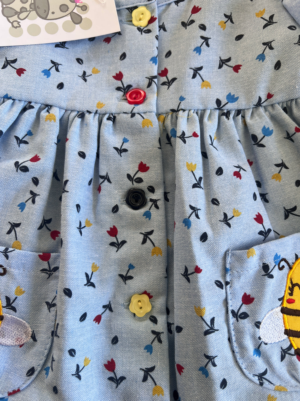Close-up of a light blue children's dress with a small floral print in red, yellow, and blue, featuring button closures and cartoon bee patch on the pocket.