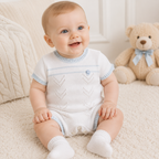 Baby wearing a white knitted romper with blue trim, sitting on a soft surface with a teddy bear in the background.