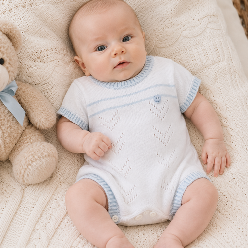 Baby in a white and blue knitted romper lying on a soft surface with a teddy bear.
