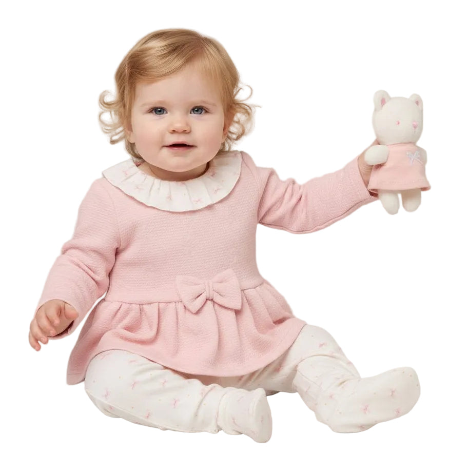 A baby girl sitting on a light gray studio background, wearing a pink ribbed knit dress with a white ruffled collar, matching pink booties, and holding a small white plush toy.