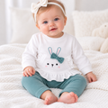 A baby girl sitting on a white textured blanket, wearing a white long-sleeve ruffle-trim top with a bunny face and teal bow applique, paired with teal pants and a white bow headband.