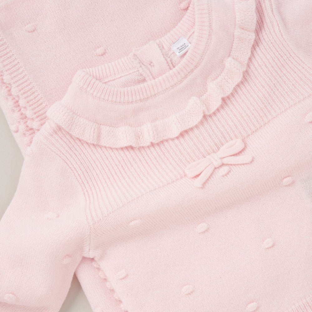 A close-up of a light pink ribbed knit baby sweater featuring a ruffled neckline, small raised dots, and a matching bow detail on the chest.