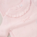A close-up of a light pink ribbed knit baby sweater featuring a ruffled neckline, small raised dots, and a matching bow detail on the chest.