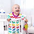 Baby in a colorful zigzag outfit standing in a crib with a toy.