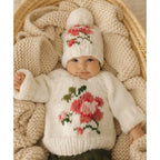 Baby wearing a knitted sweater and hat with floral patterns, lying on a textured blanket.