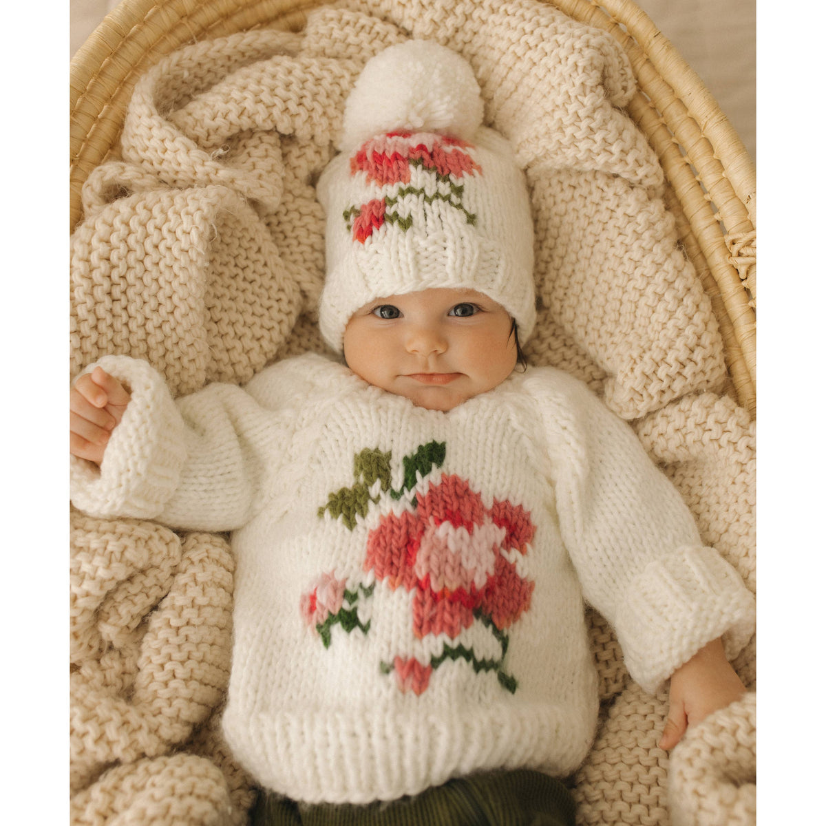 Baby wearing a knitted sweater and hat with floral patterns, lying on a textured blanket.