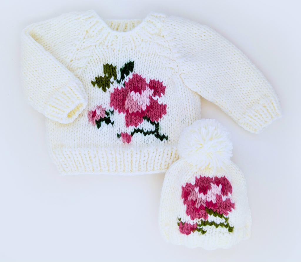 A cream knit sweater and matching knit hat displayed on a white background, both featuring a pink and green floral motif. AdoraBaby.co.uk