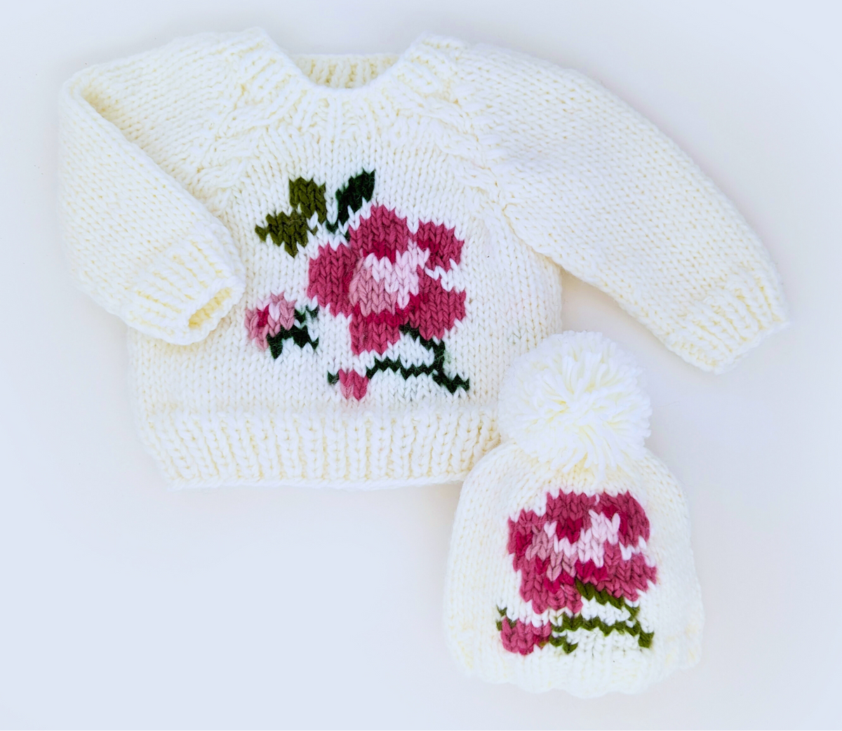 A cream knit sweater and matching knit hat displayed on a white background, both featuring a pink and green floral motif. AdoraBaby.co.uk
