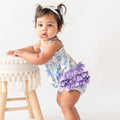 Baby girl in a floral romper with purple ruffles standing next to a small stool.