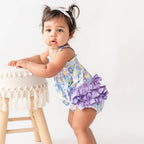Baby girl in a floral romper with purple ruffles standing next to a small stool.