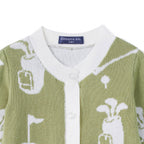 Close-up of a knitted cardigan sweater in sage green and white, featuring golf club and flag motifs, a white ribbed collar, and a visible Swaddle AN brand label.