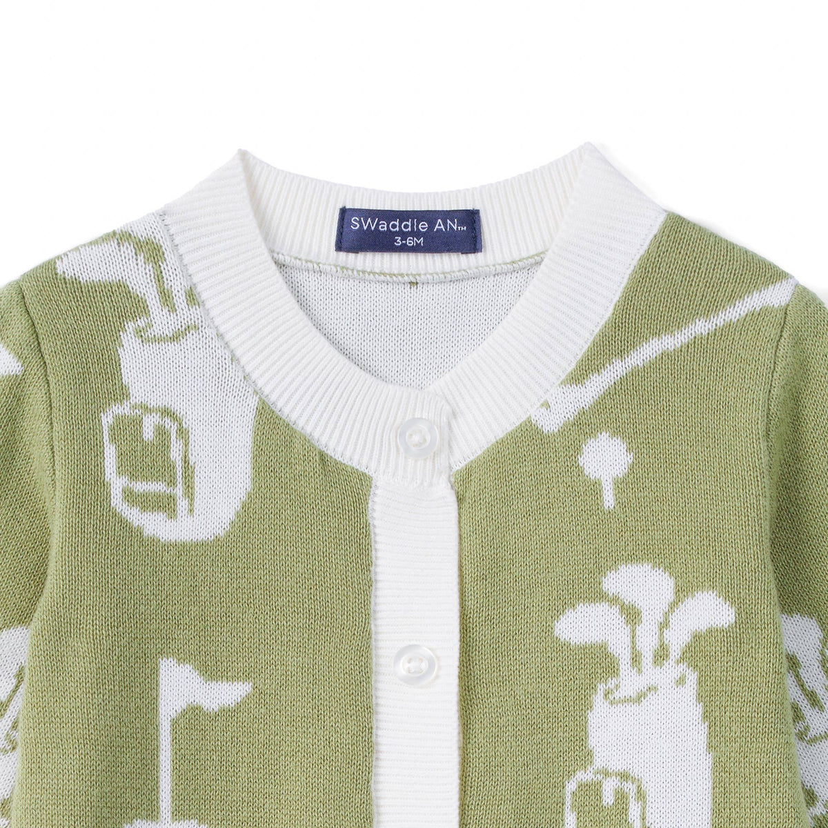 Close-up of a knitted cardigan sweater in sage green and white, featuring golf club and flag motifs, a white ribbed collar, and a visible Swaddle AN brand label.