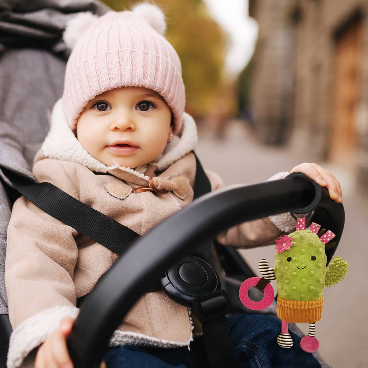Child in a stroller with a cactus toy, wearing a pink hat and beige coat.