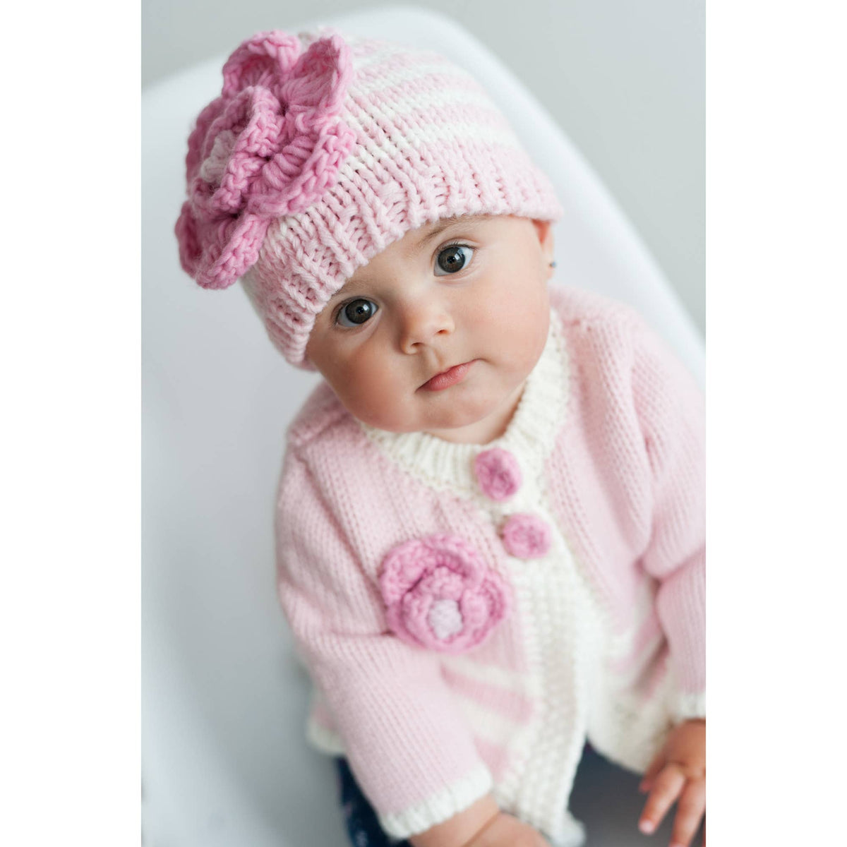 Baby wearing a pink knitted hat and cardigan with flower details on a white background