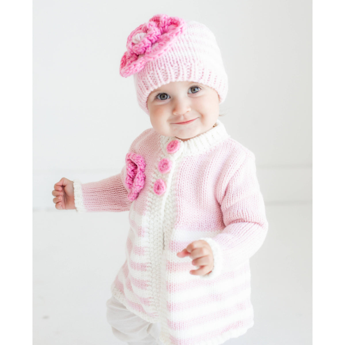 Baby wearing a pink knitted outfit with a matching hat on a white background