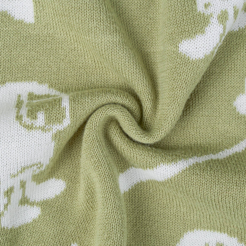 A close-up of a knitted fabric in a muted sage green color with a white knit pattern of stylized cartoon characters, arranged in a central swirl.