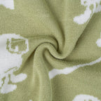 A close-up of a knitted fabric in a muted sage green color with a white knit pattern of stylized cartoon characters, arranged in a central swirl.