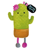 A green cactus-shaped plush toy with a textured minky fabric body, a pink flower on its head, a pink and white polka-dot bow, an orange corduroy skirt, and striped legs. A black hangtag on the right reads 'Squish Snug Cactus'.