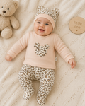A baby lying on a cream knit blanket, wearing a light pink long-sleeve top with a leopard-print heart on the chest, matching leopard-print footed pants, and a leopard-print beanie with cat ears. A beige teddy bear sits to the left, and a round wooden sign reading 'little one' is to the right.