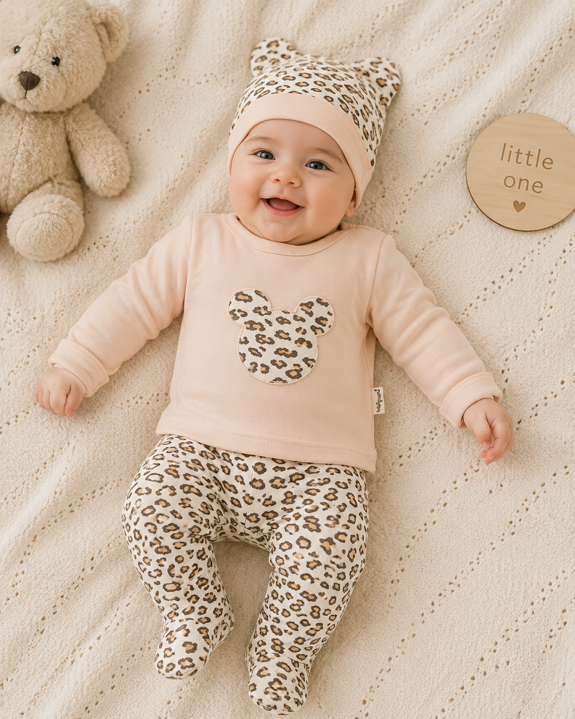 A baby lying on a cream knit blanket, wearing a light pink long-sleeve top with a leopard-print heart on the chest, matching leopard-print footed pants, and a leopard-print beanie with cat ears. A beige teddy bear sits to the left, and a round wooden sign reading 'little one' is to the right.