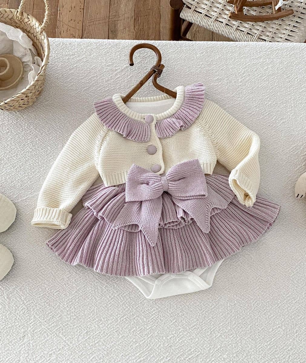 A baby girl's two-piece knit romper set featuring a cream cardigan top with a lavender pleated collar and button, paired with a matching lavender pleated ruffled skirt and large bow, displayed on a wooden hanger.