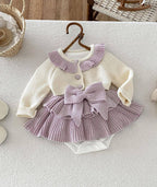 A baby girl's two-piece knit romper set featuring a cream cardigan top with a lavender pleated collar and button, paired with a matching lavender pleated ruffled skirt and large bow, displayed on a wooden hanger.