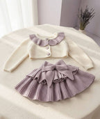 A baby girl's two-piece knit outfit set consisting of a cream cardigan with a lavender ruffled collar and a matching lavender pleated skirt with a large bow, displayed on a cream fabric-covered table.