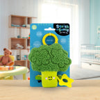 Green broccoli-shaped teether with a smiling face on a blue packaging card, set against a blurred indoor background.