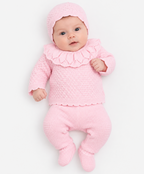 A baby wearing a soft pink knit three-piece outfit with a bonnet, ruffled collar top, and footed pants, photographed on a clean white background. AdoraBaby.co.uk