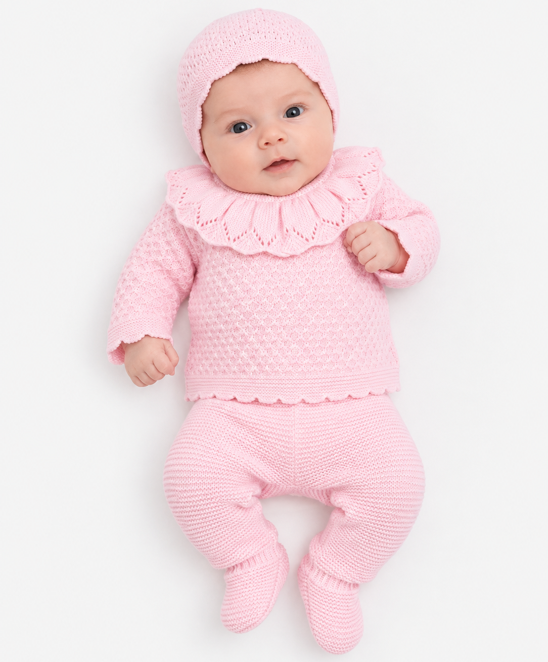 A baby wearing a soft pink knit three-piece outfit with a bonnet, ruffled collar top, and footed pants, photographed on a clean white background. AdoraBaby.co.uk