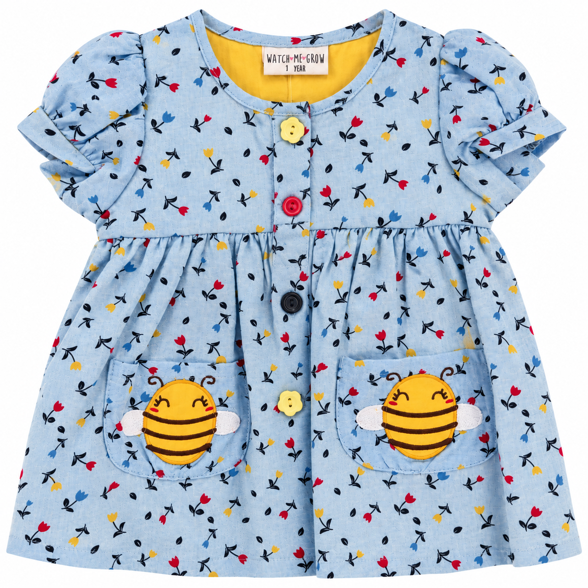 A light blue baby dress with a colorful floral print, featuring two front pockets with applique bee designs and a yellow inner collar. The brand name 'WATCH-ME-GROW' is visible on the inner neck label.